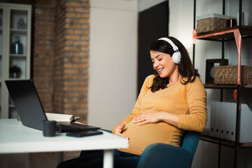 eautiful pregnant listening the music while working on laptop. Young businesswoman working in her office