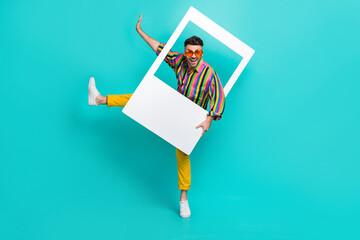 Full length photo of positive man dressed striped shirt yellow pants dancing in photo frame isolated on turquoise color background