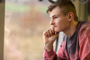 Fototapeta na wymiar Side view of young man in pink sweatshirt traveling by train, looks out of the window. Blond man enjoying a train trip.