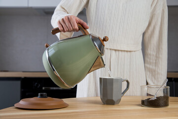 woman prepares tea in the kitchen on a wooden table. adding tea leaves to a cup and pouring hot water with an electric kettle