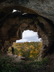 Woman standing by lighted exit in cave, daytime. Atmospheric snapshot in natural rock formations with autumn forest and dramatic gray clouds, tourist attraction. Smotrych river canyon. Vertical photo