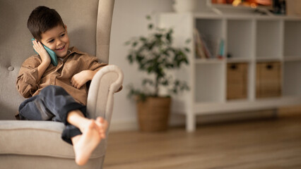 Smiling small european boy sit on armchair calling by phone, talking to parents in room interior, panorama