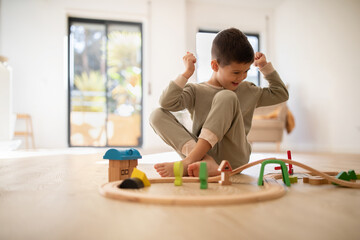 Happy small european boy in pajamas sits on floor, plays with road, rejoices in victory, success, enjoy spare time