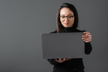 smiling asian woman in black turtleneck and glasses holding laptop isolated on dark grey