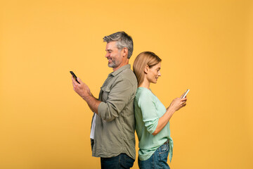 Middle aged caucasian man and woman standing back to back, using smartphones, yellow studio background, side view