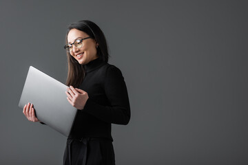 positive asian woman in black turtleneck and glasses holding laptop isolated on dark grey