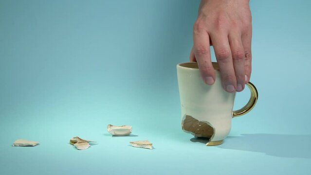 Man holding broken tea cup isolated on blue background. Close up of arm holding cracked coffee mug