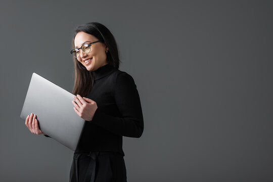 Positive Asian Woman In Black Turtleneck And Glasses Holding Laptop Isolated On Dark Grey