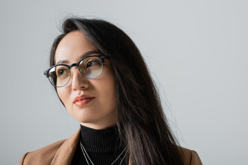 portrait of brunette asian businesswoman in beige blazer and glasses looking away isolated on grey