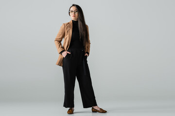 full length of confident asian woman in beige blazer and black pants standing with hands in pockets...