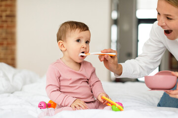 Breakfast for kid. Mother feeding her adorable baby daughter with porridge, sitting on bed in bedroom interior, free space