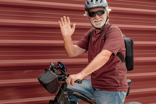 Handsome senior man riding his electric bicycle in the city wearing helmet and sunglasses standing against red metal background looking at camera and waving