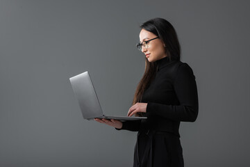pretty asian woman in black turtleneck and glasses using laptop isolated on dark grey