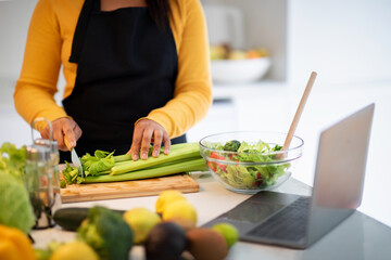 Cheerful millennial african american lady in apron making salad, cuts celery at table with laptop