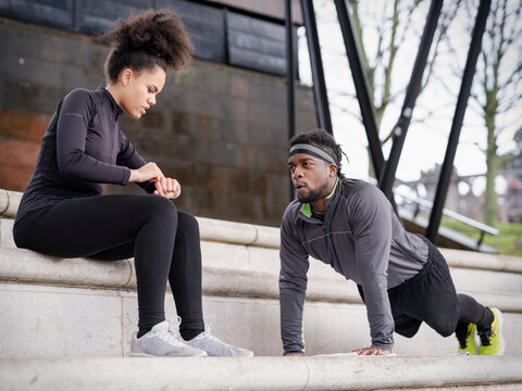 Athletic woman assisting man doing push-ups on steps outdoors