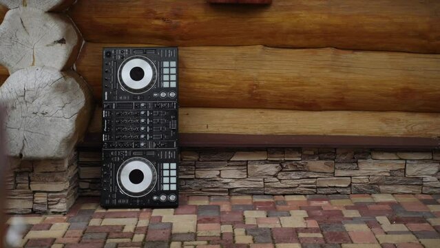 Party DJ mixing console lies upright on outdoor tiles, leaning against house of thick wooden logs. Digital professional audio equipment.