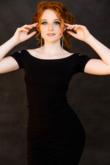 Beautiful slender red-haired girl with straight earrings in a tight dress posing on a black background.