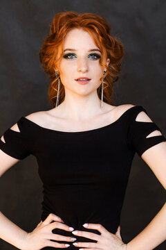 Beautiful slender red-haired girl with straight earrings in a tight dress posing on a black background.