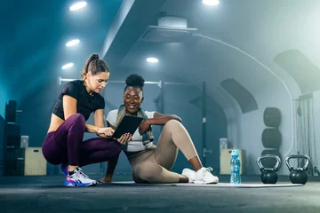 Schilderijen op glas Fitness gym instructor showing workout plan on tablet to young African American woman in sportswear with towel. They are sitting on the gym floor, resting after workout. © MexChriss