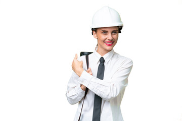 Young architect woman with helmet and holding blueprints over isolated background pointing back