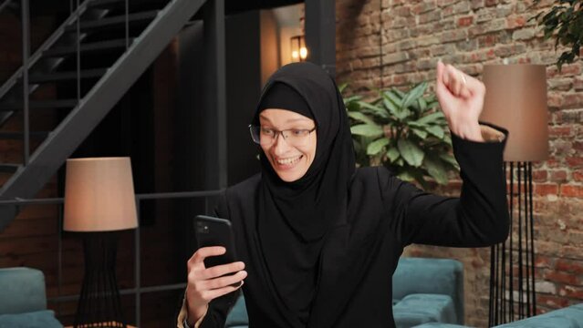 Surprised Muslim Islamic woman winner hold smartphone read good news feels euphoric amazed by mobile online bet bid game win winner, overjoyed by victory success. Financial stock sports betting.