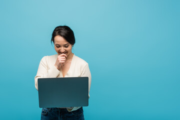 Excited asian woman in cardigan laughing and holding laptop isolated on blue