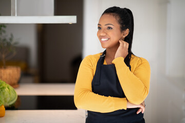 Cheerful pretty millennial african american lady chef in apron cooking food in white modern kitchen interior