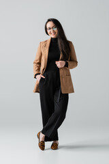 full length of happy asian businesswoman in beige blazer and black pants standing on grey