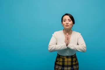 Asian woman in cardigan showing praying hands gesture isolated on blue