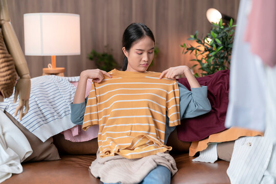 Uncertain young asian woman in casual clothes sitting on comfortable couch trying to decide between cloth  against others in living room at home,tired exhausted girl makeing decision for her cloth