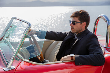 young man in convertible car looking at camera, smiling, man wears suit and tie drives red convertible car