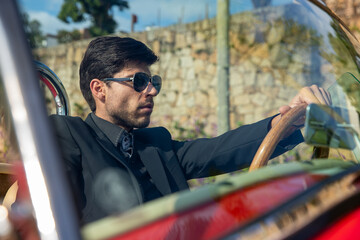 young man in convertible car looking at camera, smiling, man wears suit and tie drives red convertible car