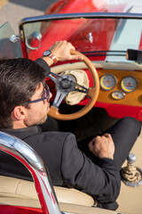 young man in convertible car looking at camera, smiling, man wears suit and tie drives red convertible carman in car
