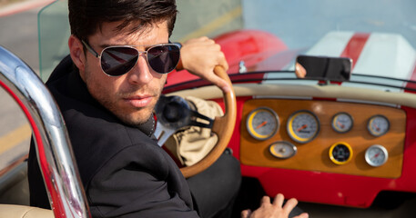 young man in convertible car looking at camera, smiling, man wears suit and tie drives red...
