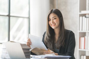 Looking camera, Beautiful Asian female bookkeeper doing online accounting with laptop in private accounting office, balance sheet and stock market statistics.