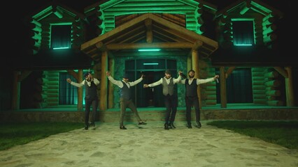 Group of young men dancing synchronized with alcohol drink bottles and glasses . Drunk dancers having fun , celebrating , dancing folk dance on classic outfits . Restaurant or house on background 