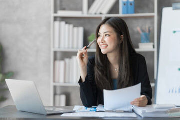 Beautiful Asian female bookkeeper doing online accounting with laptop in private accounting office, balance sheet and stock market statistics.