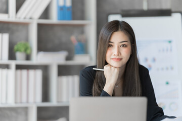 Looking camera, Beautiful Asian female bookkeeper doing online accounting with laptop in private accounting office, balance sheet and stock market statistics.