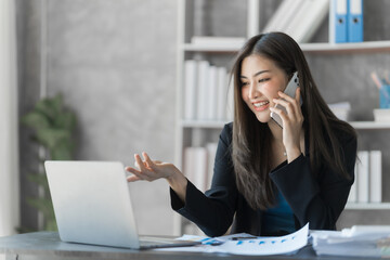 Call mobile, using laptop, Beautiful Asian female bookkeeper doing online accounting with laptop in private accounting office, balance sheet and stock market statistics.