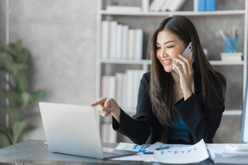 Call mobile, using laptop, Beautiful Asian female bookkeeper doing online accounting with laptop in private accounting office, balance sheet and stock market statistics.