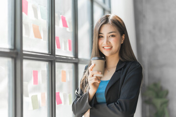 Holding coffee up and looking camera, Pretty asia business woman bookkeeper brainstorming and working in modern office workplace with sticky notes or post it at windows.