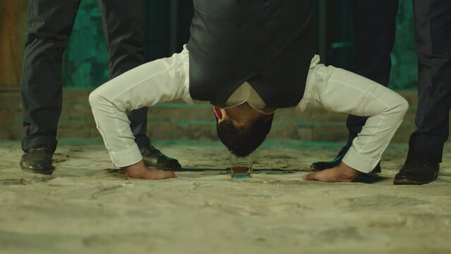 Young man flipping upside down on the floor , doing trick with glass of alcohol drink , drinking . His friends applauding cheering him . Men celebrating , having fun, laughing . Drinking without hands