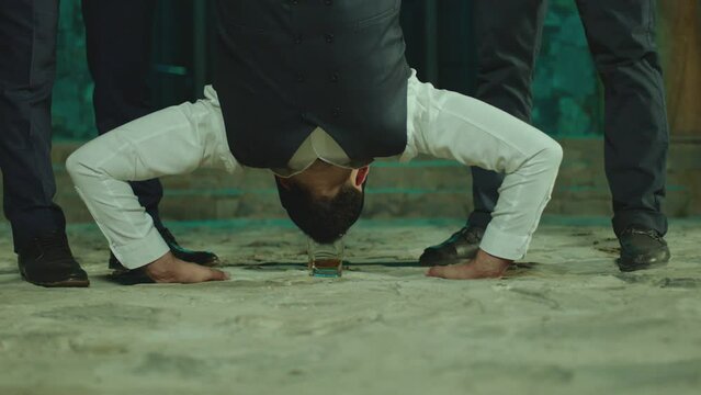 Young man flipping upside down on the floor , doing trick with glass of alcohol drink , drinking . His friends applauding cheering him . Men celebrating , having fun, laughing . Drinking without hands