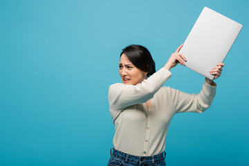 Aggressive asian freelancer throwing laptop isolated on blue