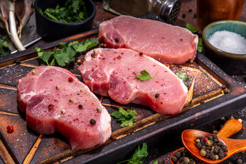 Raw pork steaks cooking background. Three tasty raw fresh meat steaks with herbs, salt, pepper, olive oil, ready for dinner preparation, dark brown kitchen background copy space