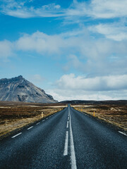 A winding, desolate road stretches out ahead, surrounded by flat, barren earth as a towering mountain range rises on the horizon, shrouded in clouds