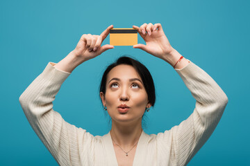 Pensive asian woman looking at credit card above head isolated on blue