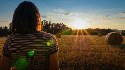 A young women in a striped t-shirt und shoulder long brown hair is looking at sunset with sunflare over a harvested field with hay bales | dreaming, freedom, hope