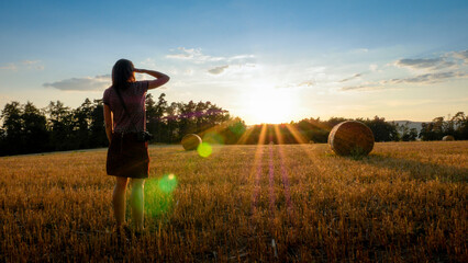 A young women is shielding her eyes and looking at  sunset with sunflare over a harvested field with hay bales.