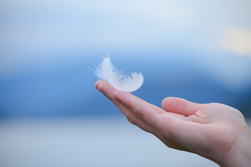 Young woman's hand holding white soft feather against morning sky, spiritual background,...